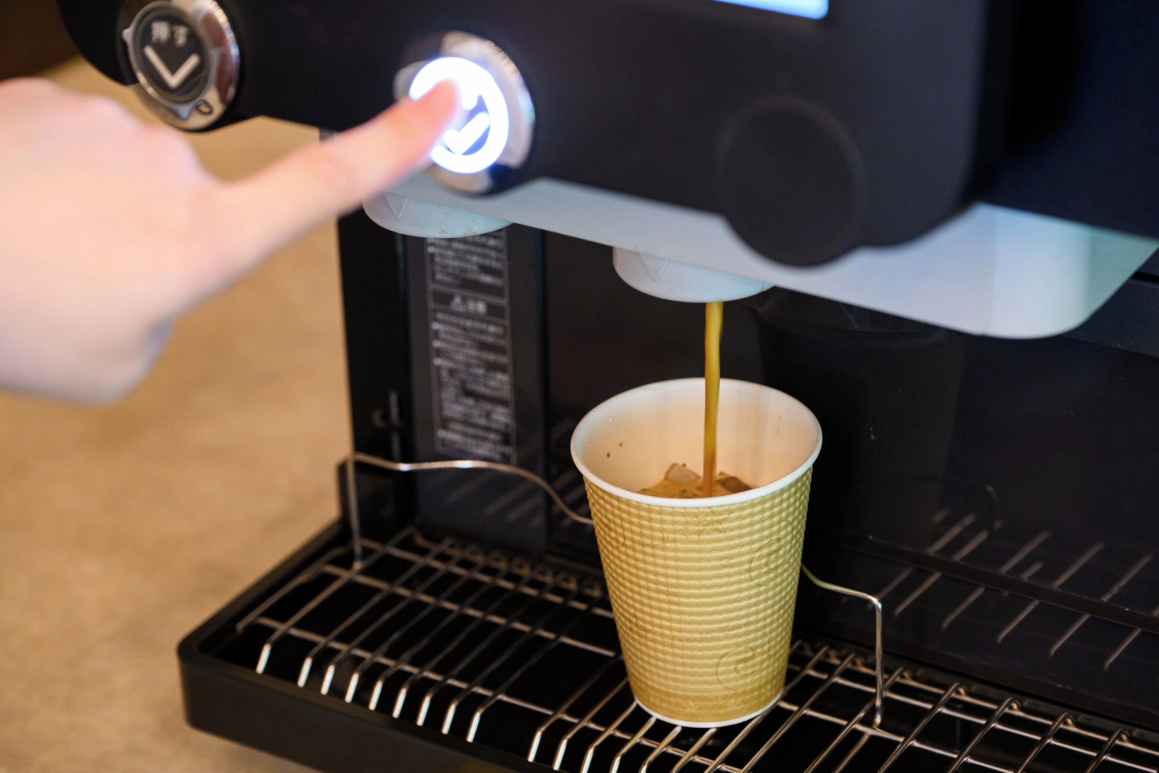Eine Hand drückt eine Taste an einer Kaffeemaschine, während Kaffee in einem Papierbecher gefüllt wird.