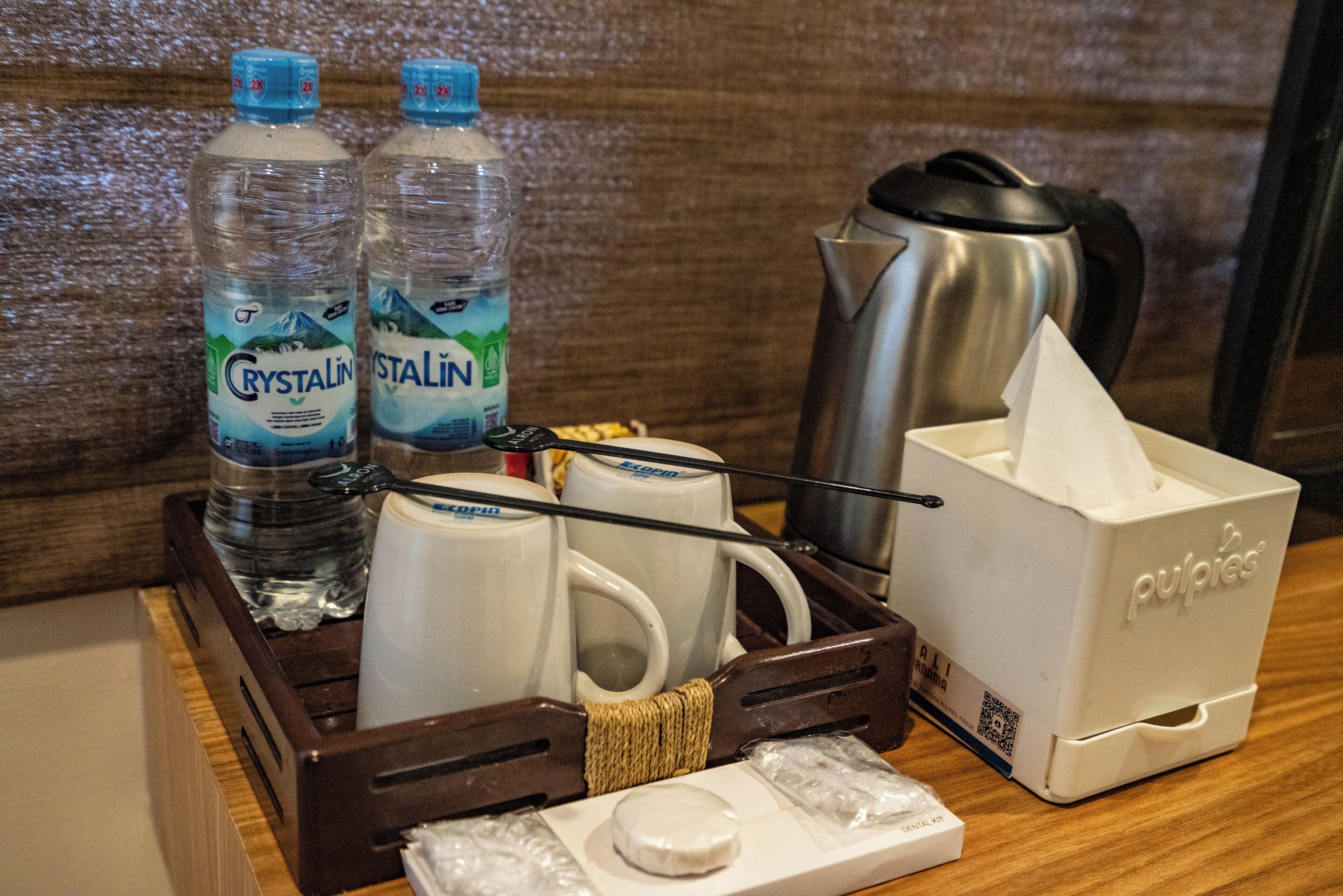 Bandeja de desayuno en una habitación de hotel, con dos botellas de agua Crystalina, dos tazas blancas, una cafetera de acero inoxidable, una caja de pañuelos y un pequeño recipiente con un objeto blanco.