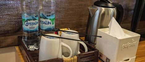 Bandeja de desayuno en una habitación de hotel, con dos botellas de agua Crystalina, dos tazas blancas, una cafetera de acero inoxidable, una caja de pañuelos y un pequeño recipiente con un objeto blanco.