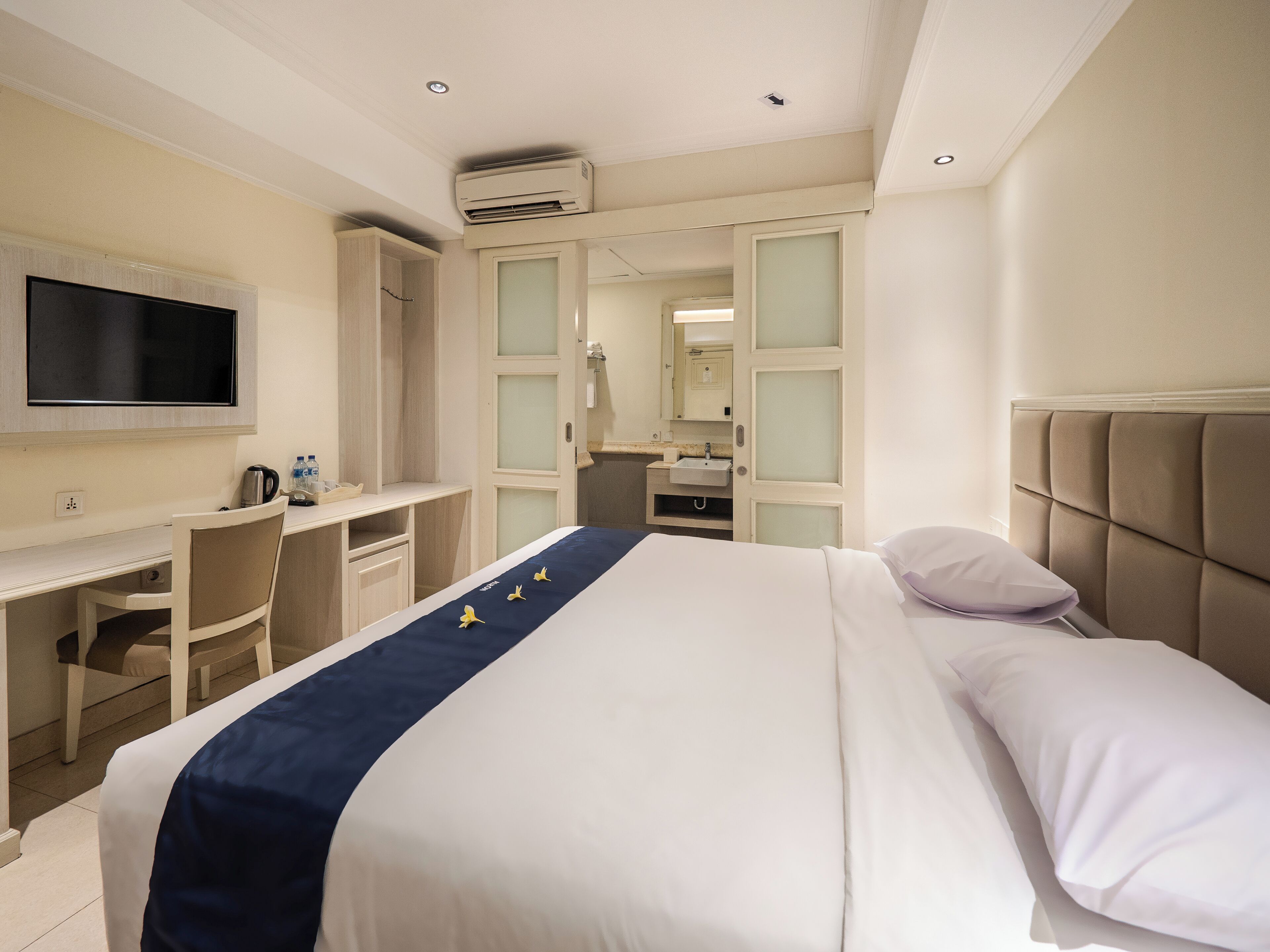 Quarto de hotel com uma cama, escrivaninha, cadeira, televisão e banheiro visível através de uma porta aberta.