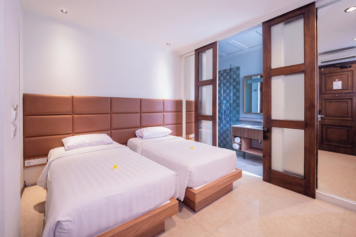 A hotel room with two beds, a wooden headboard, a bathroom with a glass shower, and a wooden wardrobe.