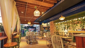 A restaurant interior with wooden ceiling beams, hanging pendant lights, and a bar area with a stone wall and greenery. - Alron Hotel Kuta Powered by Archipelago (Kuta)