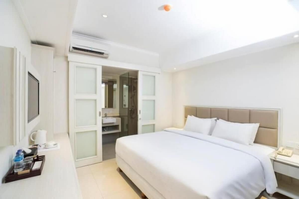 A modern hotel room with a large bed, a television, a desk with a coffee maker, and a bathroom with a glass shower enclosure.