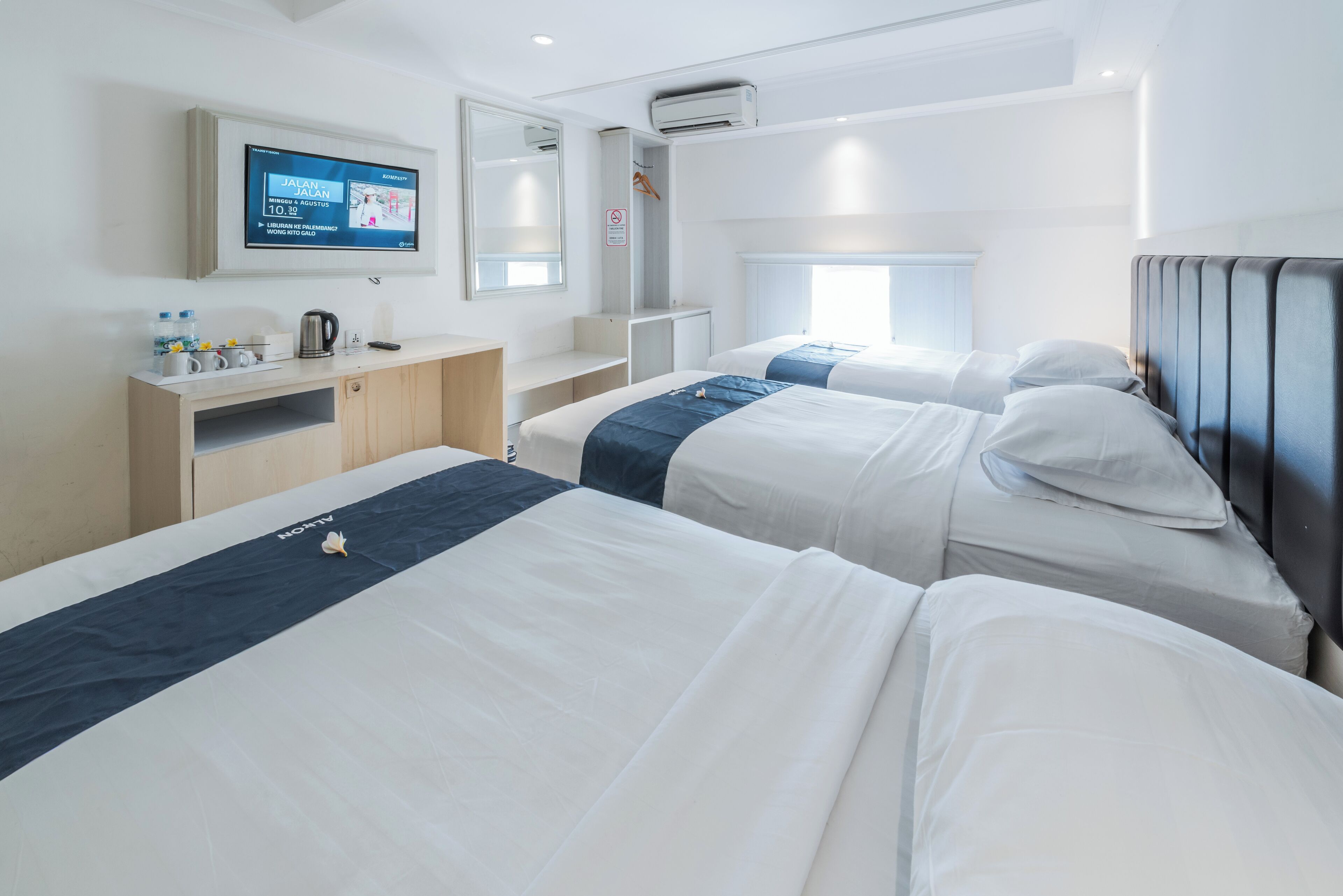 A hotel room with two beds, a TV mounted on the wall, a small desk with a kettle, and a window with a view.