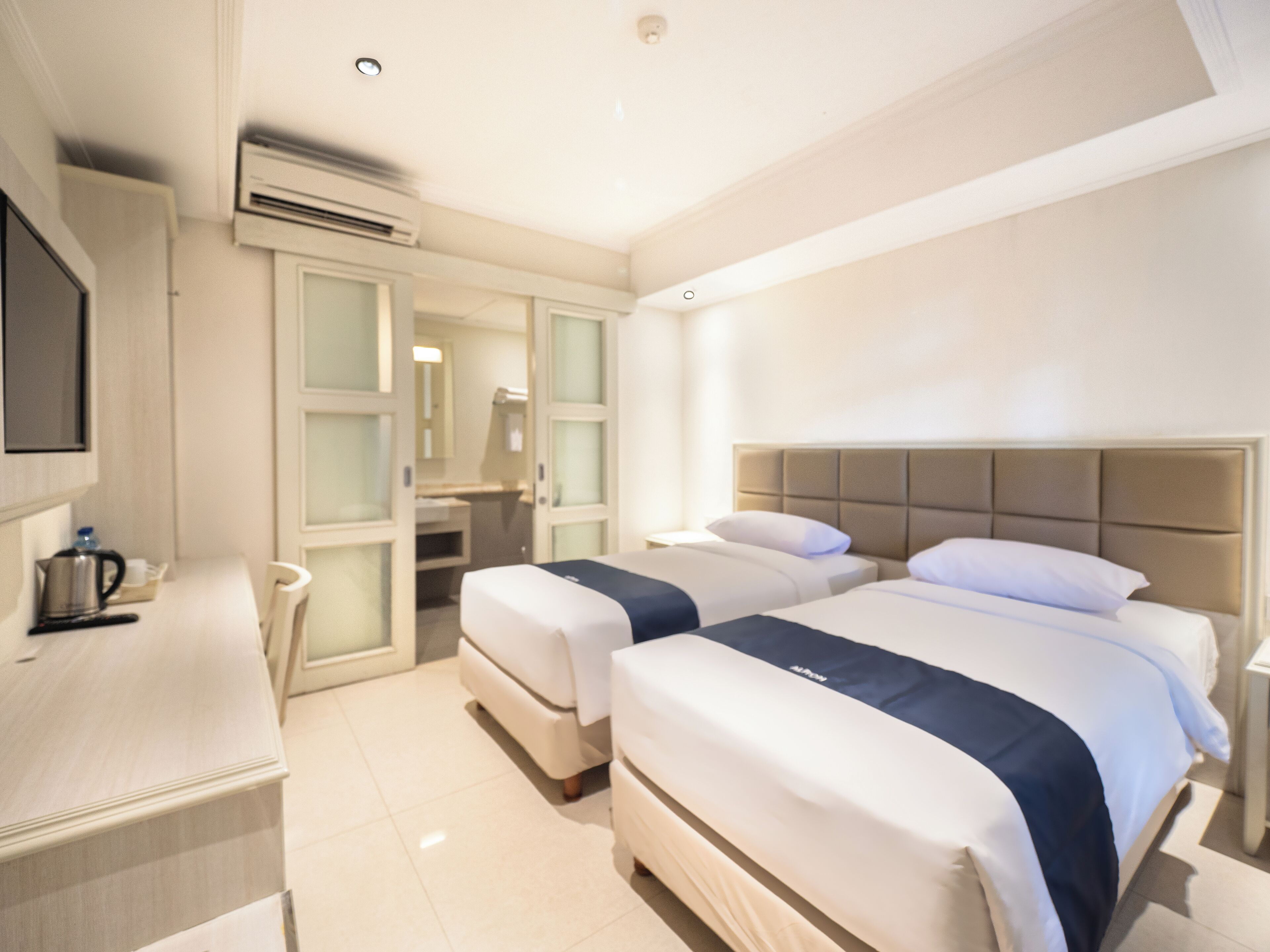 A hotel room with two beds, a desk, a mirror, and an air conditioning unit.