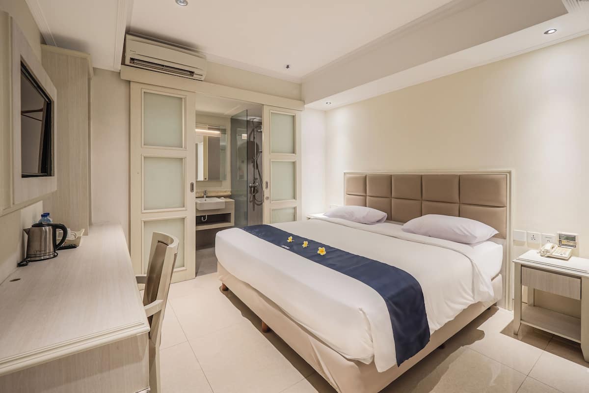 A hotel room with a bed, a desk, a chair, a nightstand, and a bathroom with a shower.