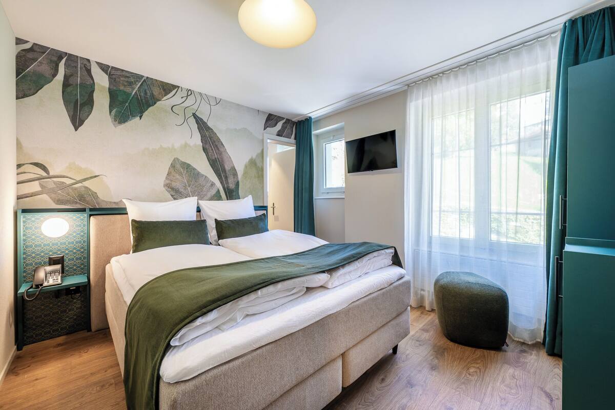 Quarto com cama, mesa de cabeceira com telefone, televisão na parede e janela com cortinas.