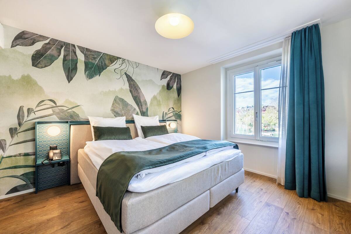 Quarto com cama, mesa de cabeceira com telefone, janela com cortinas e parede com padrão de folhas.