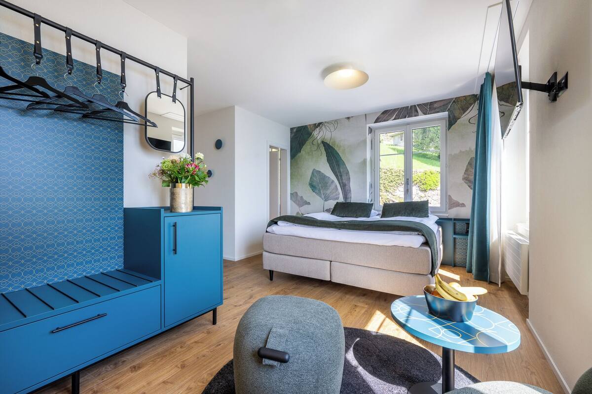 Quarto moderno com cama, sofá azul, mesinha com uma tigela de fruta e janela com vista para o verde.
