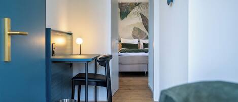 A compact room with a desk, chair, and a bed. There is a trash bin and a wall with a leaf pattern.