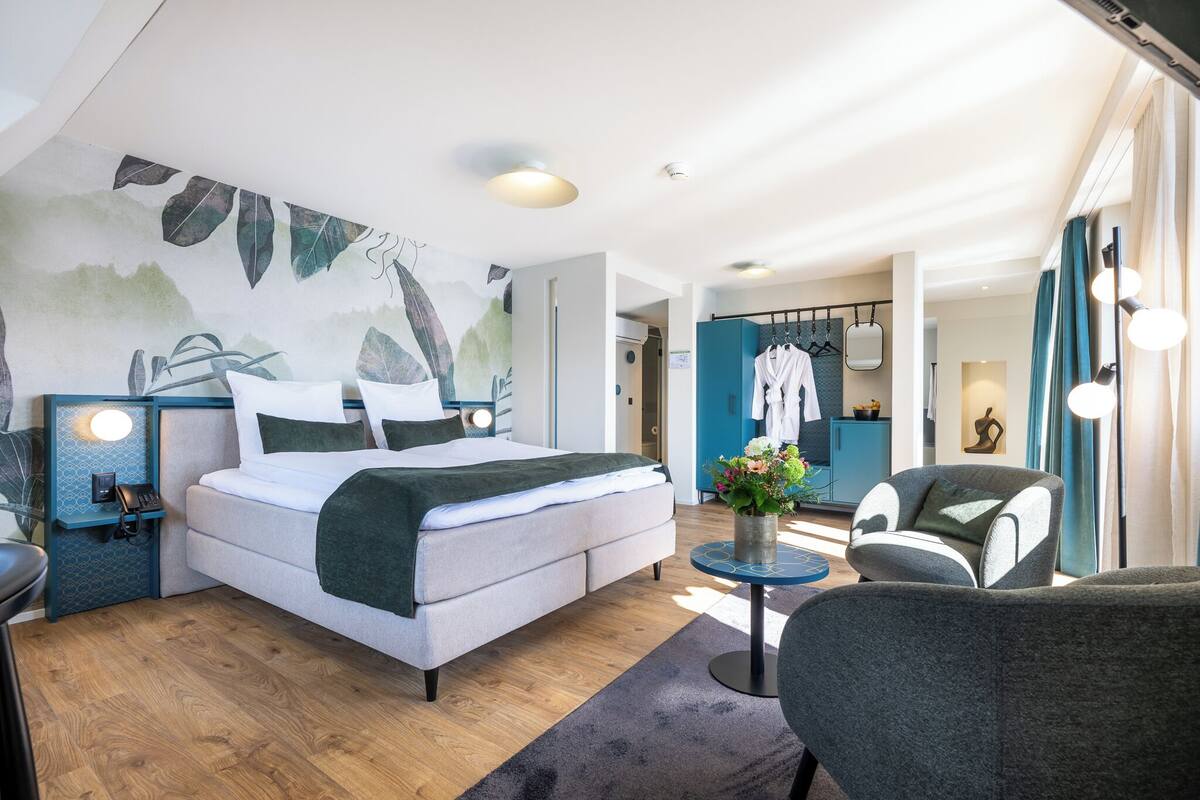 Quarto de hotel moderno com cama, área de estar, toucador com espelho e uma parede com mural temático da natureza.