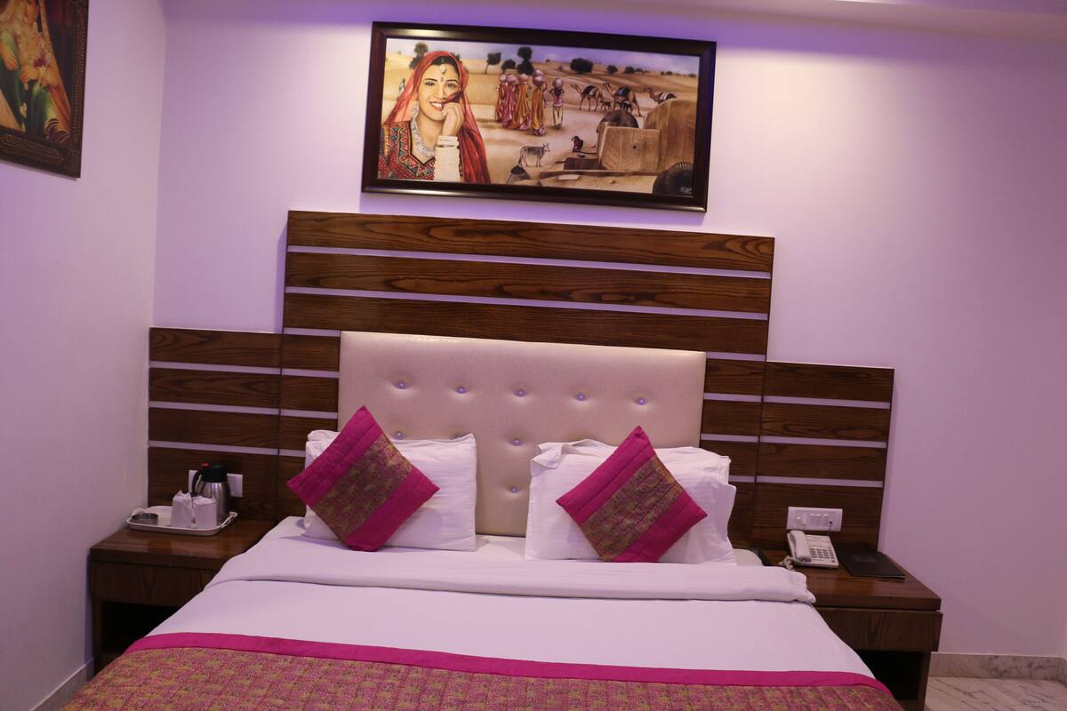 A hotel room with a large bed, wooden headboard, two pink pillows, a painting above the bed, and a nightstand with a phone and a tray.