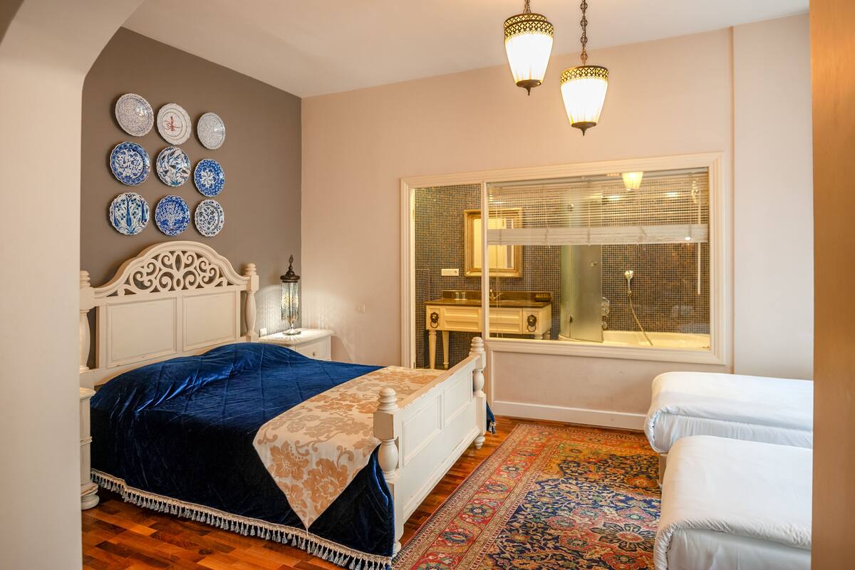 Un dormitorio con cama, mesitas de noche, una ventana con persianas y una alfombra estampada.