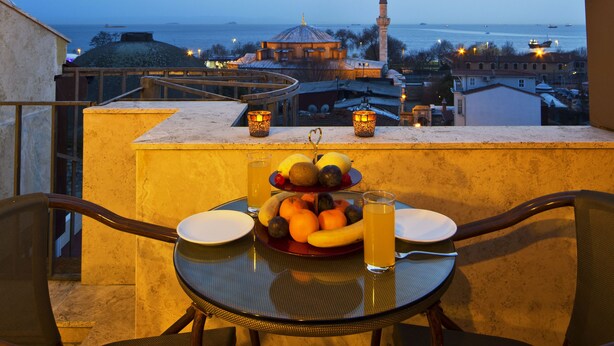Una mesa con fruta y un vaso de jugo en un balcón con vista a una ciudad, una mezquita y barcos en el agua.