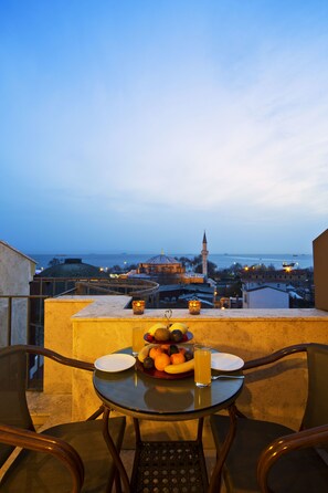 Une table avec des fruits et un verre de jus sur un balcon donnant sur un paysage urbain, avec une mosquée et des bateaux sur l’eau.
