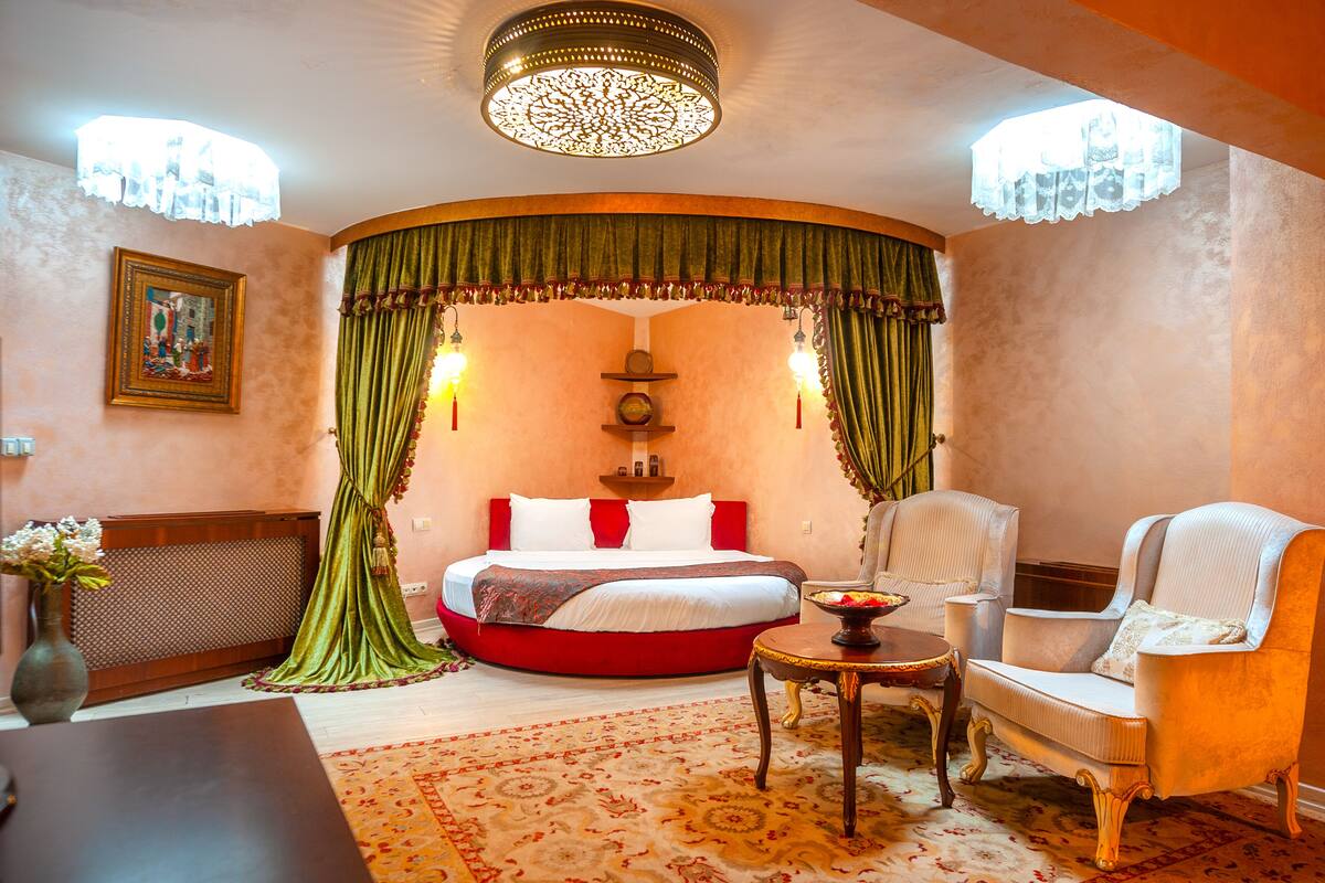 Un dormitorio con una cama curvada, cortinas verdes, un candelabro, una mesita de noche y dos sillas.