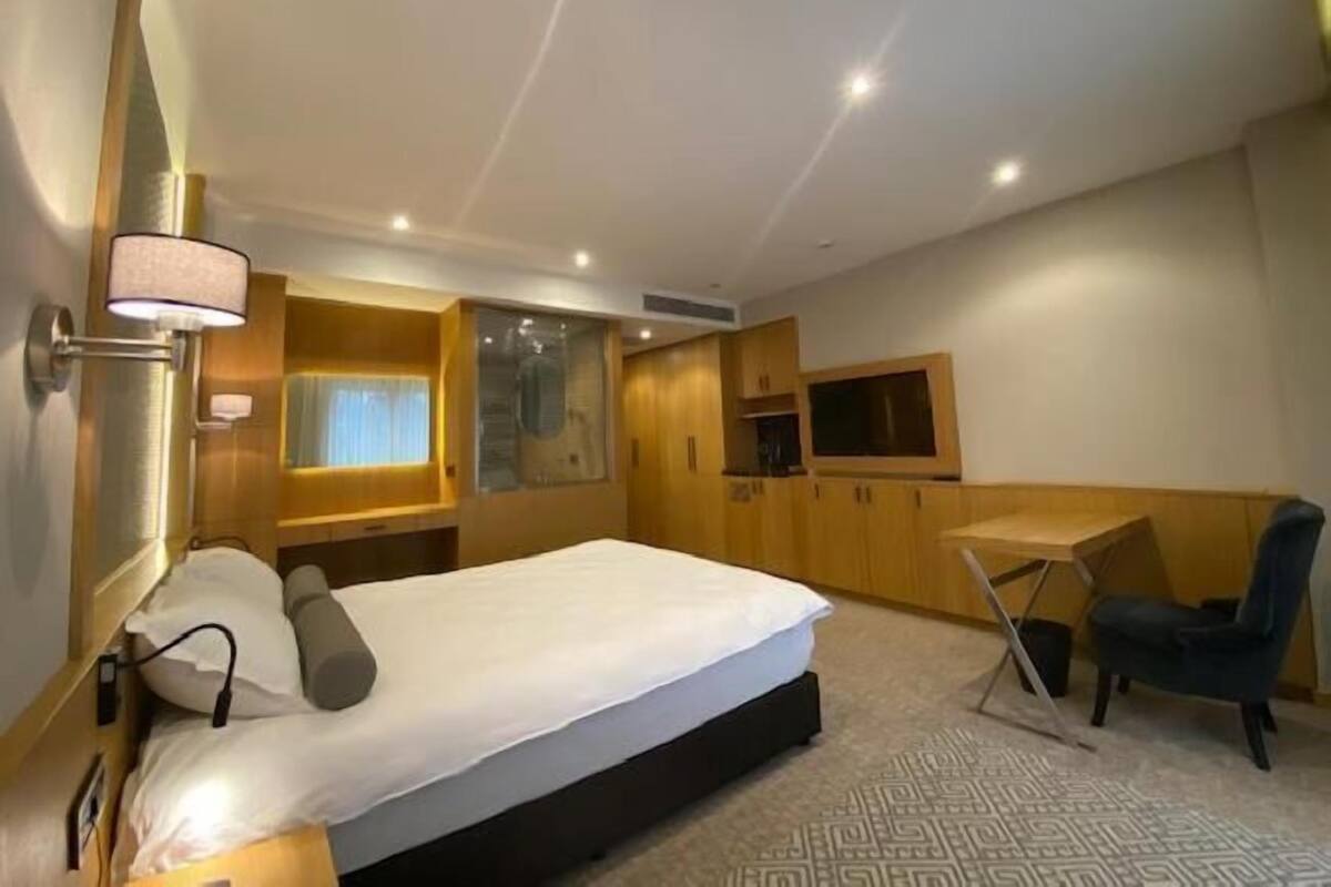 A hotel room with a bed, bedside table, lamp, and a small desk with a chair.