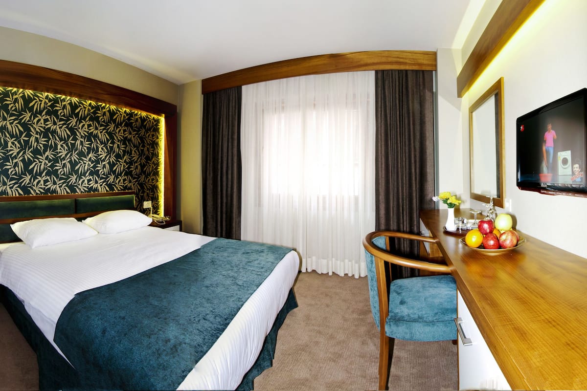 A hotel room with a large bed, a desk with a chair, a mirror, and a wall-mounted TV.