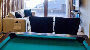 Une table de billard recouverte d’un drap vert, un canapé avec des coussins, deux fauteuils, un téléviseur posé sur un meuble et une fenêtre avec des rideaux.