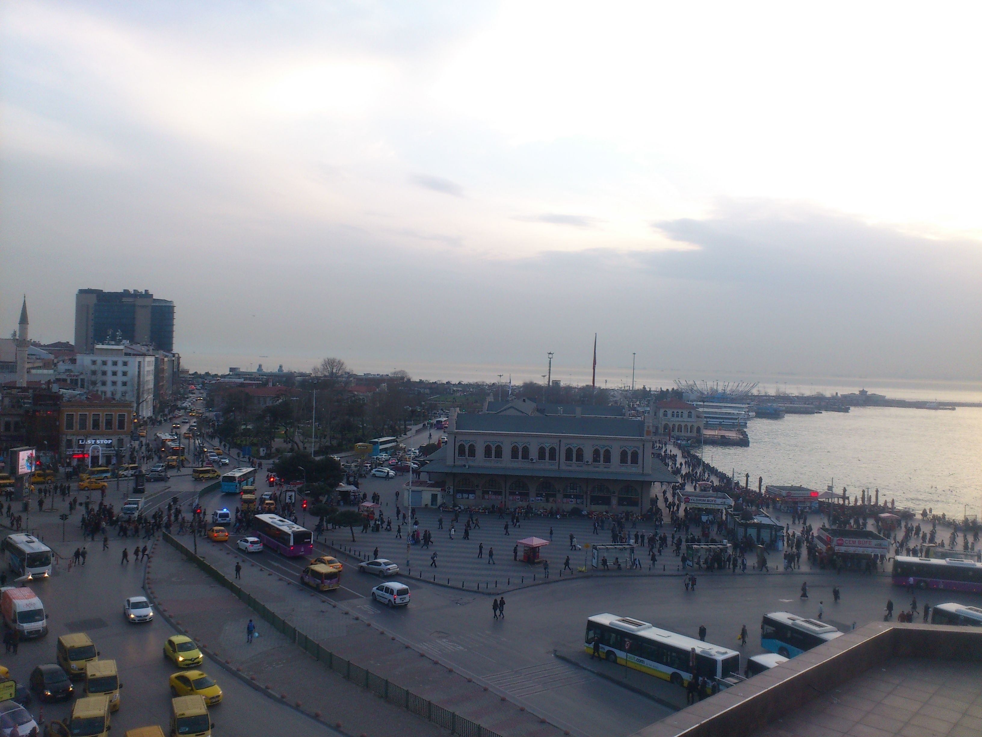 A busy urban waterfront with a pedestrian promenade, buses, and a variety of buildings including a prominent mosque.