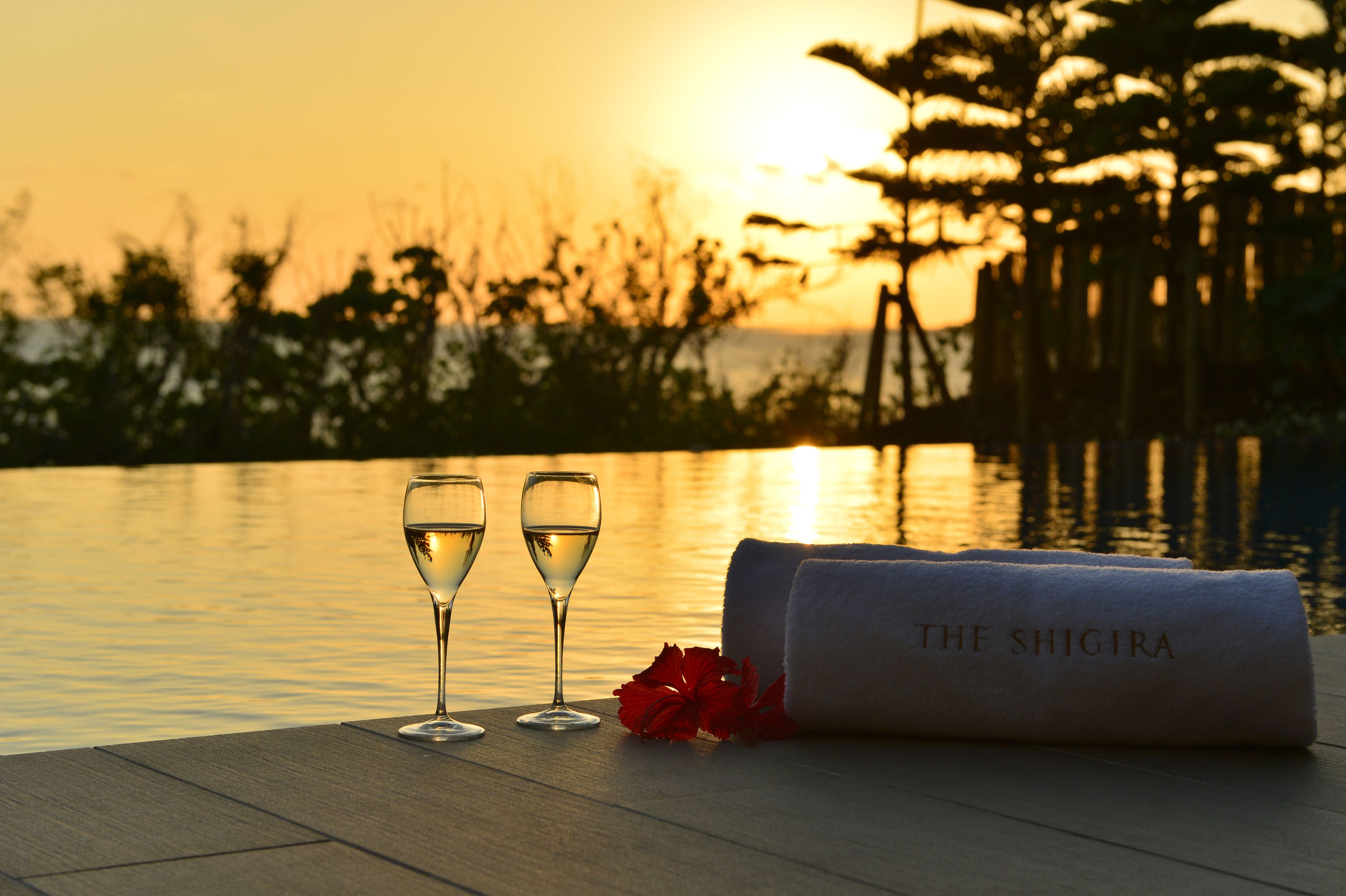 A sunset view with two glasses of white wine and a towel labeled "The Shigira" on a wooden deck by a body of water.