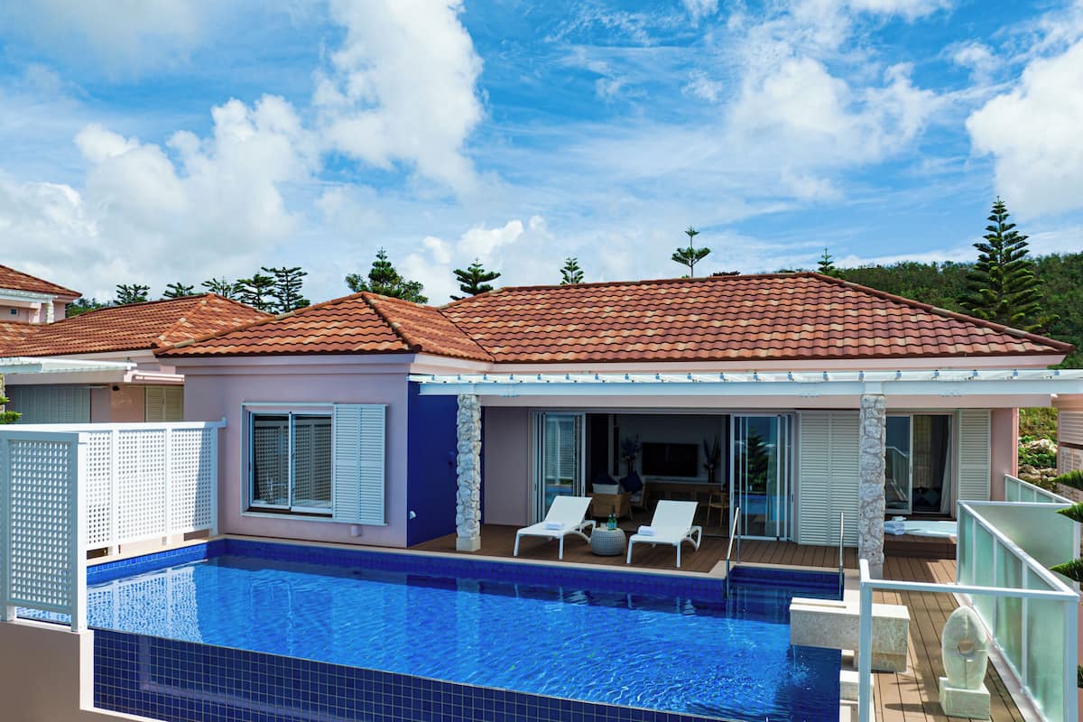 A poolside area with a clear blue pool, a covered patio with lounge chairs, and a house with a tiled roof.