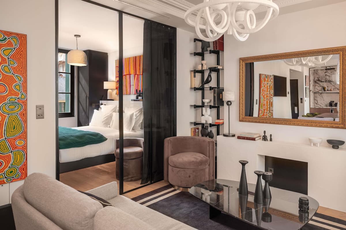 A modern hotel room with a glass partition, a sofa, a chair, a fireplace, and a large mirror.