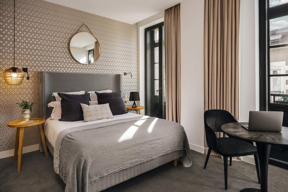 A modern hotel room with a large bed, a round table with a laptop, a small side table, and a large window with curtains.