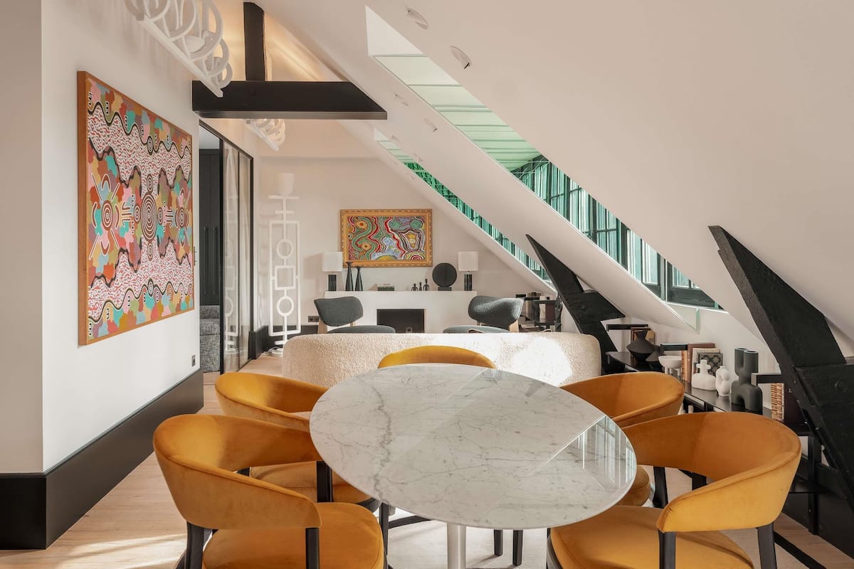 A modern interior with a round marble table, yellow chairs, and a staircase with a glass railing.