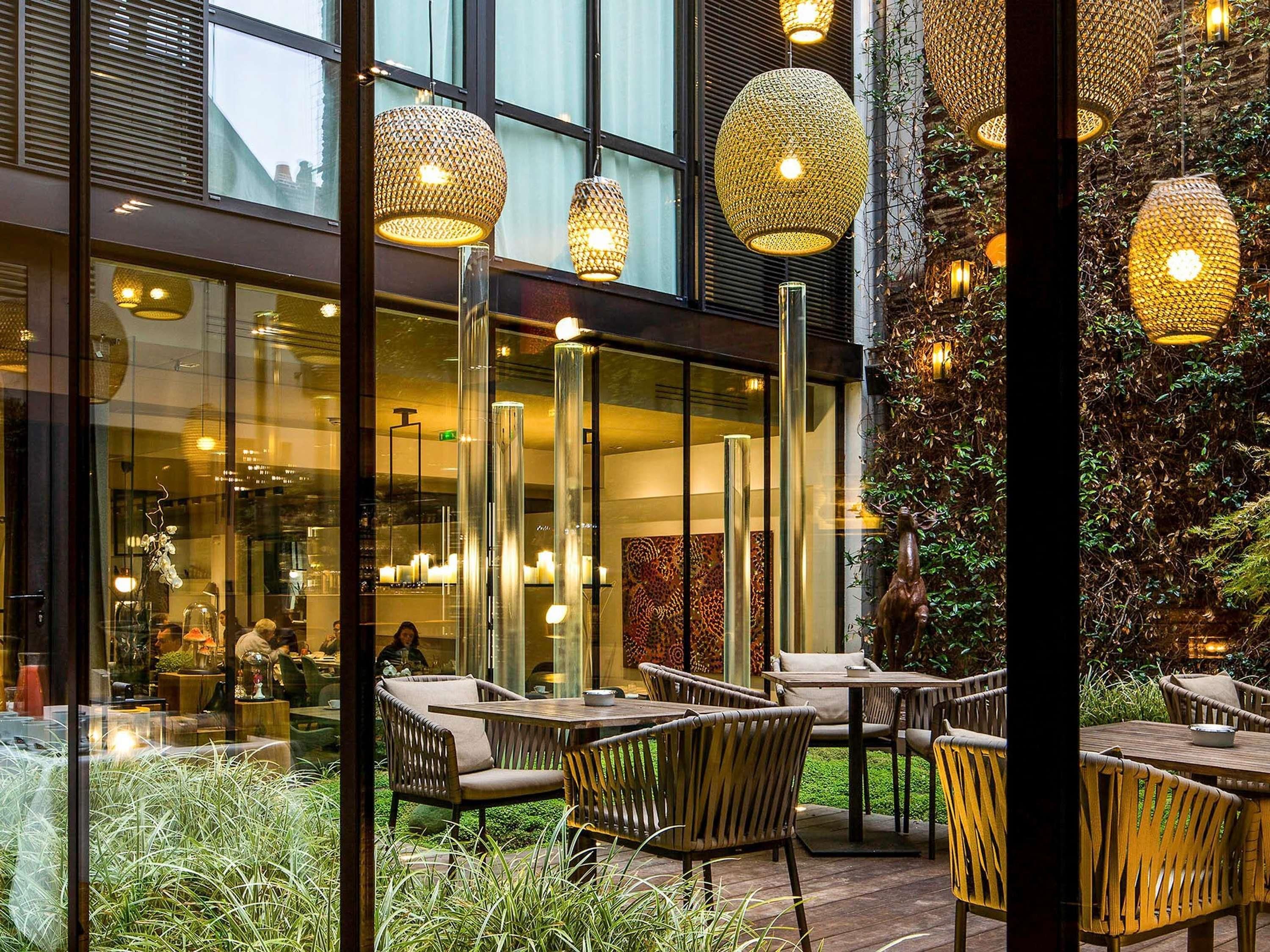 An outdoor seating area with wicker chairs and tables, surrounded by greenery and a glass wall reflecting the interior.