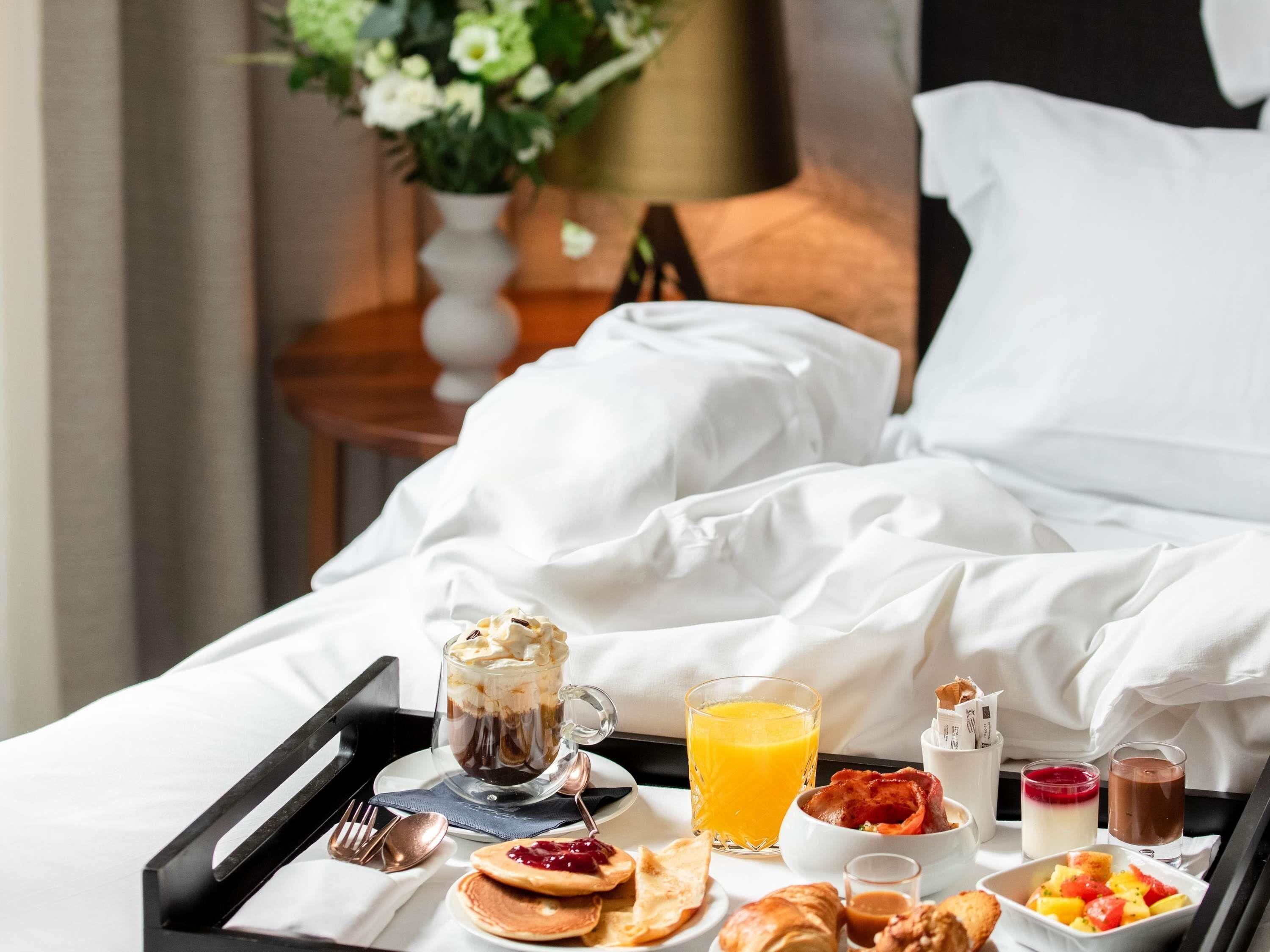 Een netjes opgemaakt ontbijtkarretje op een bed, met een stapel pannenkoeken, een glas sinaasappelsap, een kop koffie, een fruitschaal en een klein potje jam.