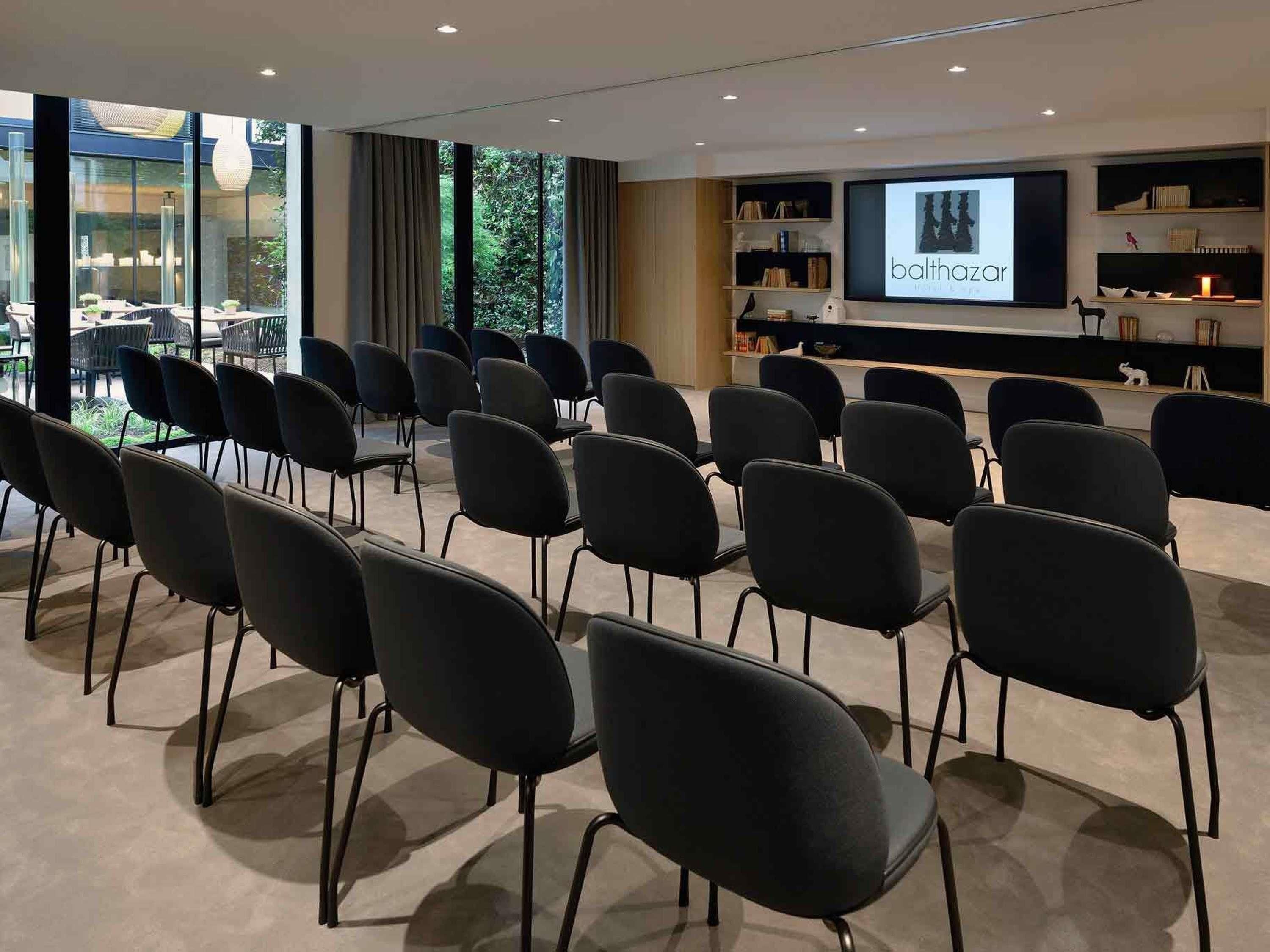 A conference room with rows of chairs facing a projection screen, a bookshelf with books and decorative items, and a view of the outdoors through large windows.