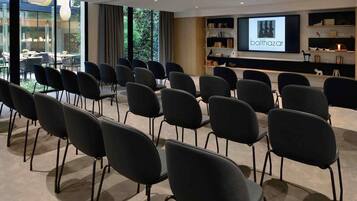 A conference room with rows of chairs facing a projection screen, a bookshelf with books and decorative items, and a view of the outdoors through large windows.