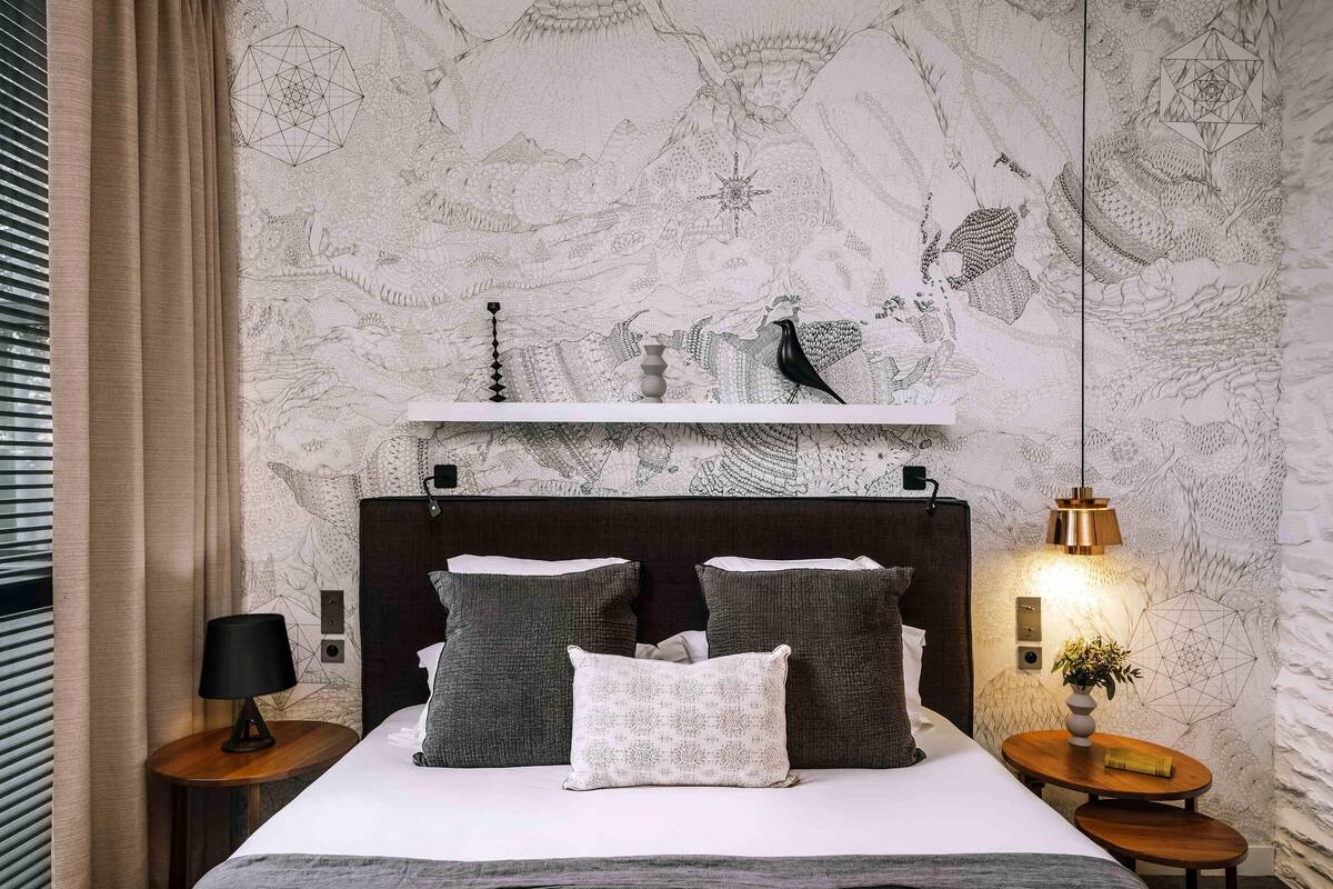 A bedroom with a large bed, a headboard, a bedside table with a lamp, and a wall with intricate patterns.