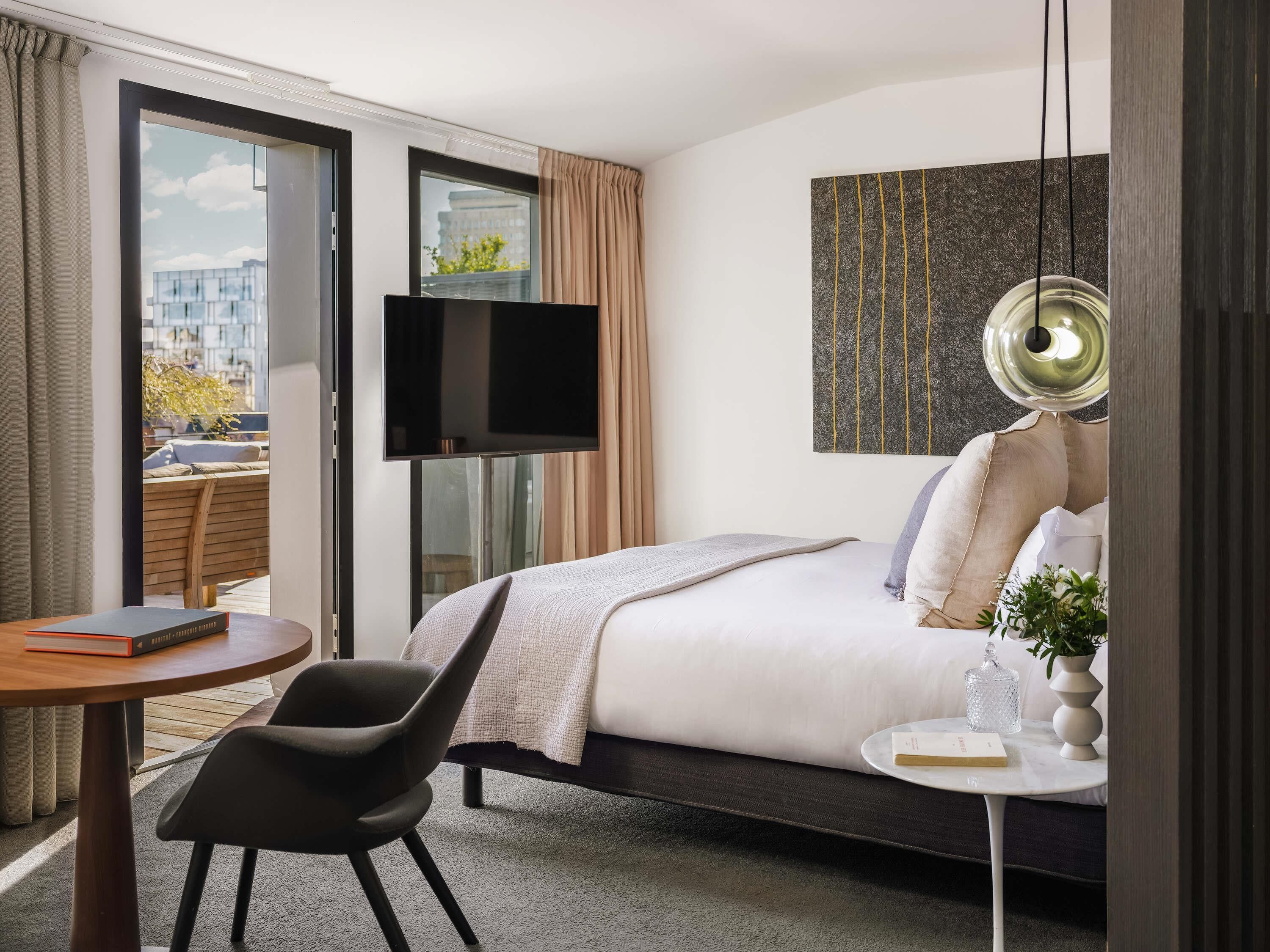 A hotel room with a bed, a TV, a round table, a chair, a small side table, and a view of buildings and greenery outside.