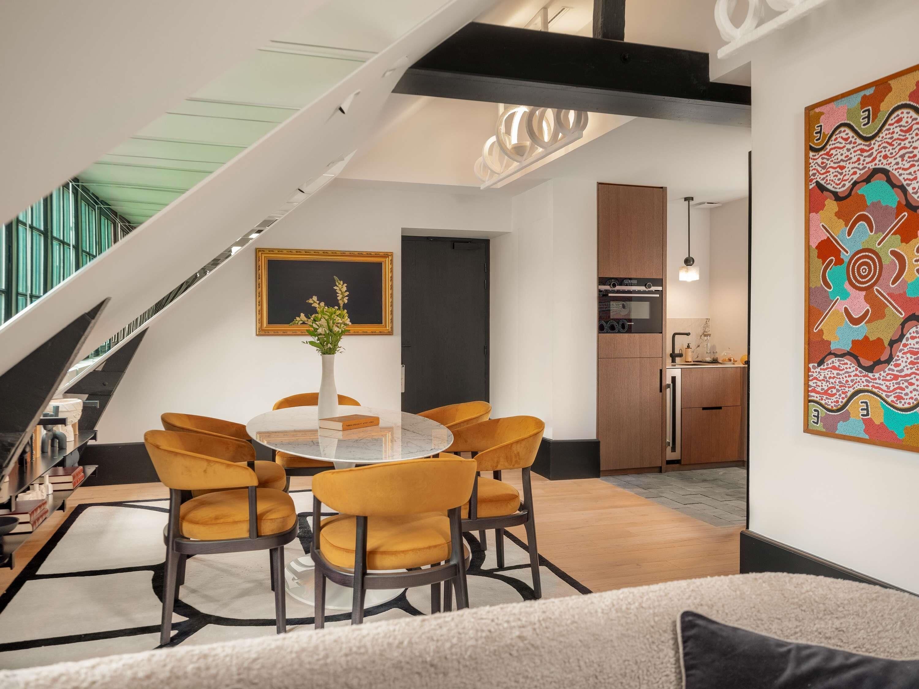 A modern living room with a round table, yellow chairs, and a sofa. There is a staircase leading to an upper level, a kitchen area, and a large abstract painting on the wall.