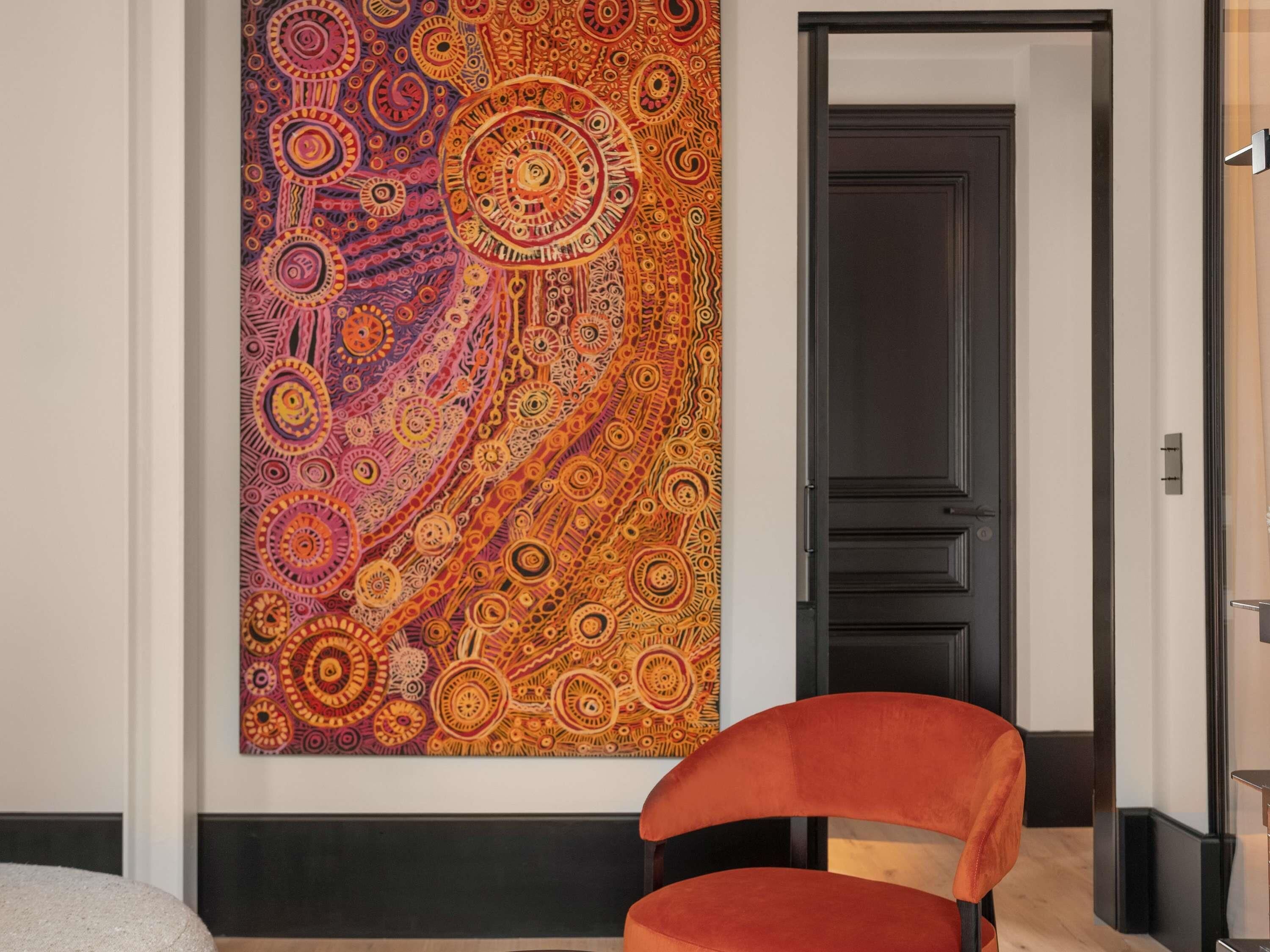 Une pièce avec une grande tapisserie abstraite au mur, une porte sombre et un fauteuil rouge.