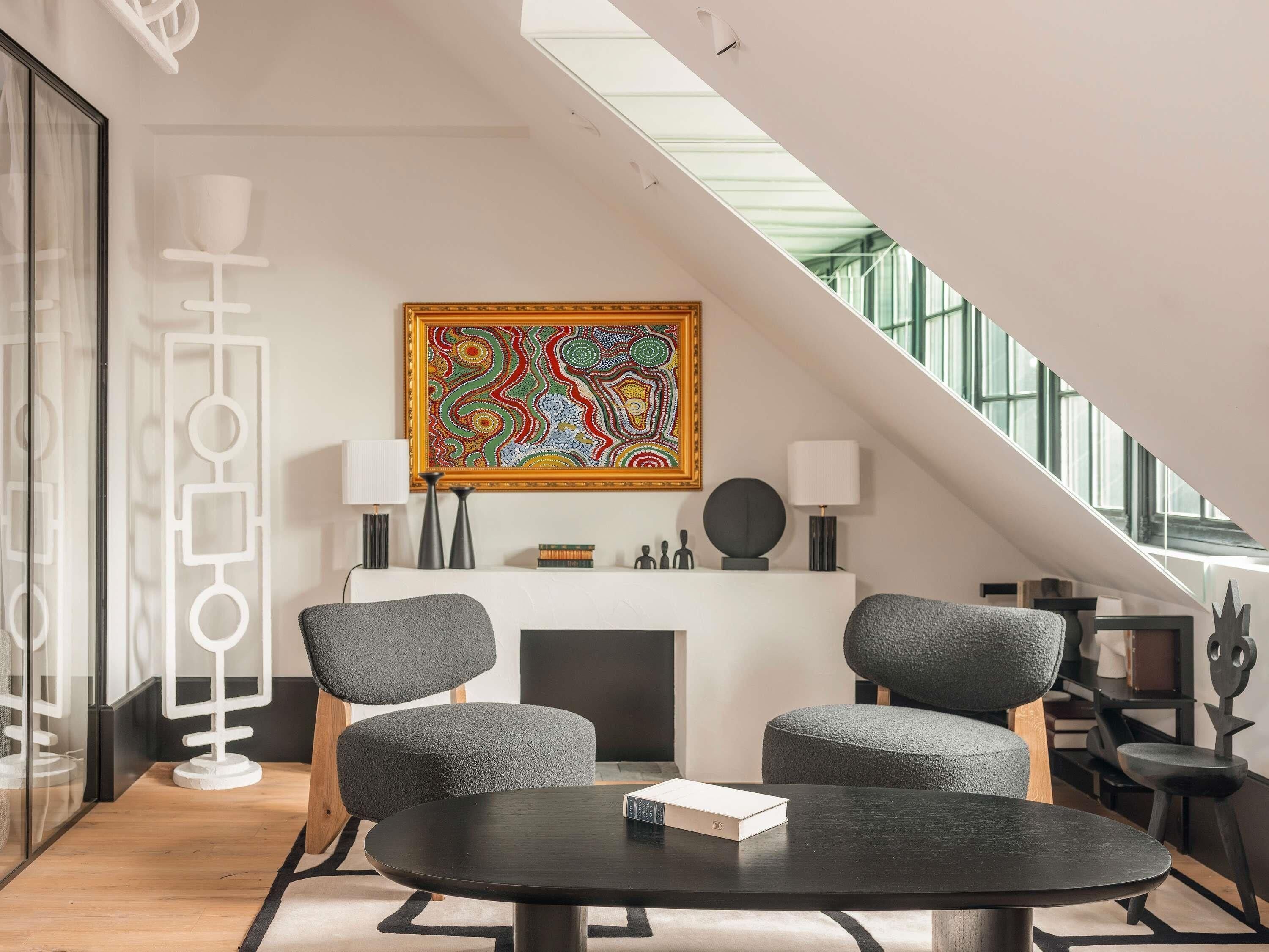 Een moderne loft met hoge plafonds, een open haard, twee grijze fauteuils, een ronde tafel en een kleurrijk abstract schilderij.