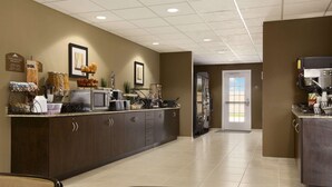 A hotel breakfast area with a long counter, various food items, and a refrigerator. - Microtel Inn & Suites by Wyndham Kenedy/Karnes City (Kenedy)