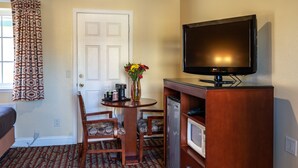 A hotel room with a TV, a small table with a vase of flowers, a sofa, patterned carpet, and a door. - Seaside Inn (Seaside)