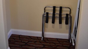 A corner of a room with a beige wall, a wooden shelf, a metal rack with black straps, and a patterned carpet. - Seaside Inn (Seaside)