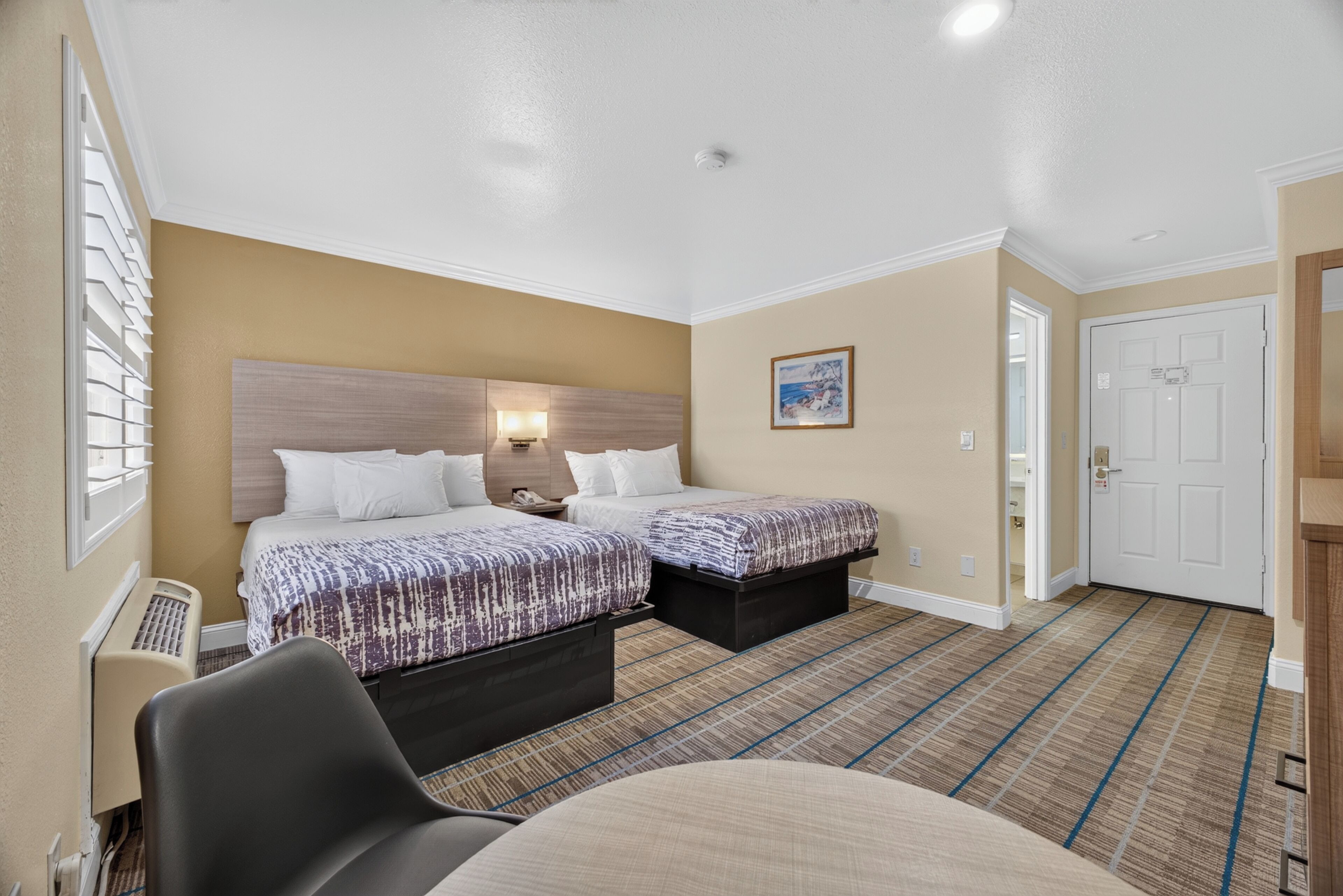 A hotel room with two beds, a desk, a chair, a radiator, and a door.