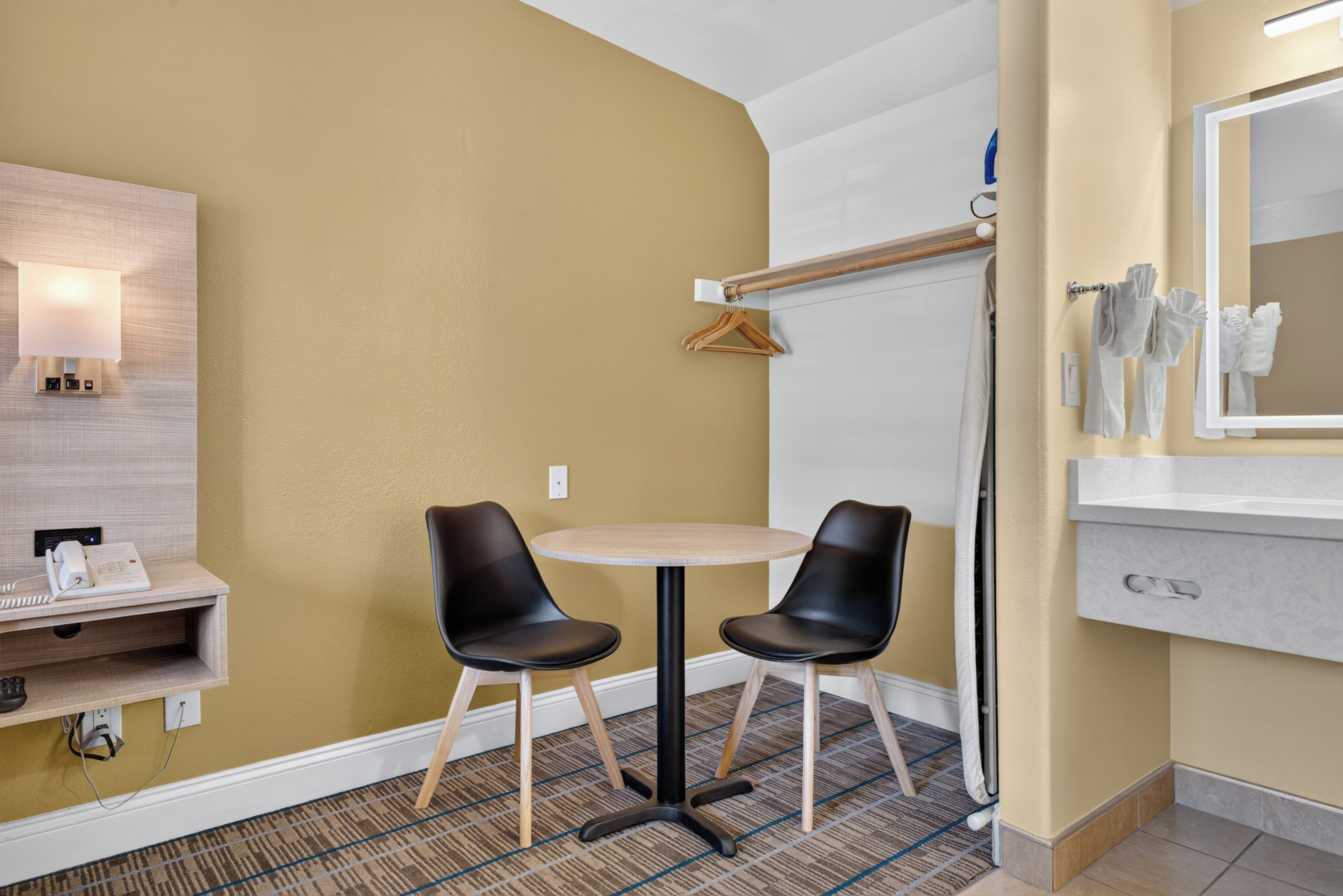 Habitación de hotel con cama, mesita de noche con lámpara, espejo en la pared, pequeña mesa redonda con dos sillas y piso alfombrado.