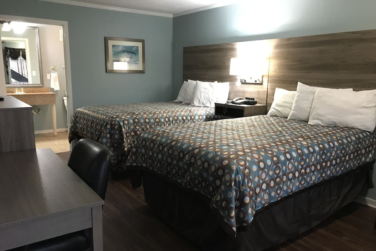 Quarto de hotel com duas camas, uma mesa e uma cadeira. É visível um banheiro por uma porta aberta.