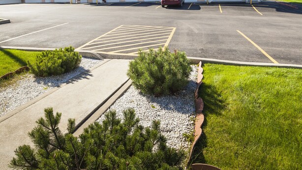 Un motel con aparcamiento, una escalera que lleva a la entrada y un pequeño jardín con arbustos y grava.