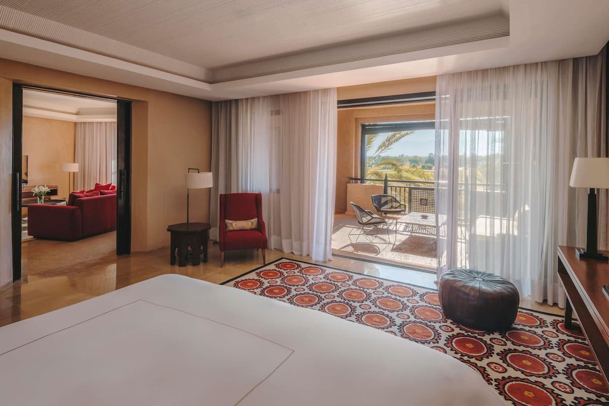 Une chambre d’hôtel avec un grand lit, un canapé rouge, une petite table, une chaise et un balcon donnant sur des palmiers.