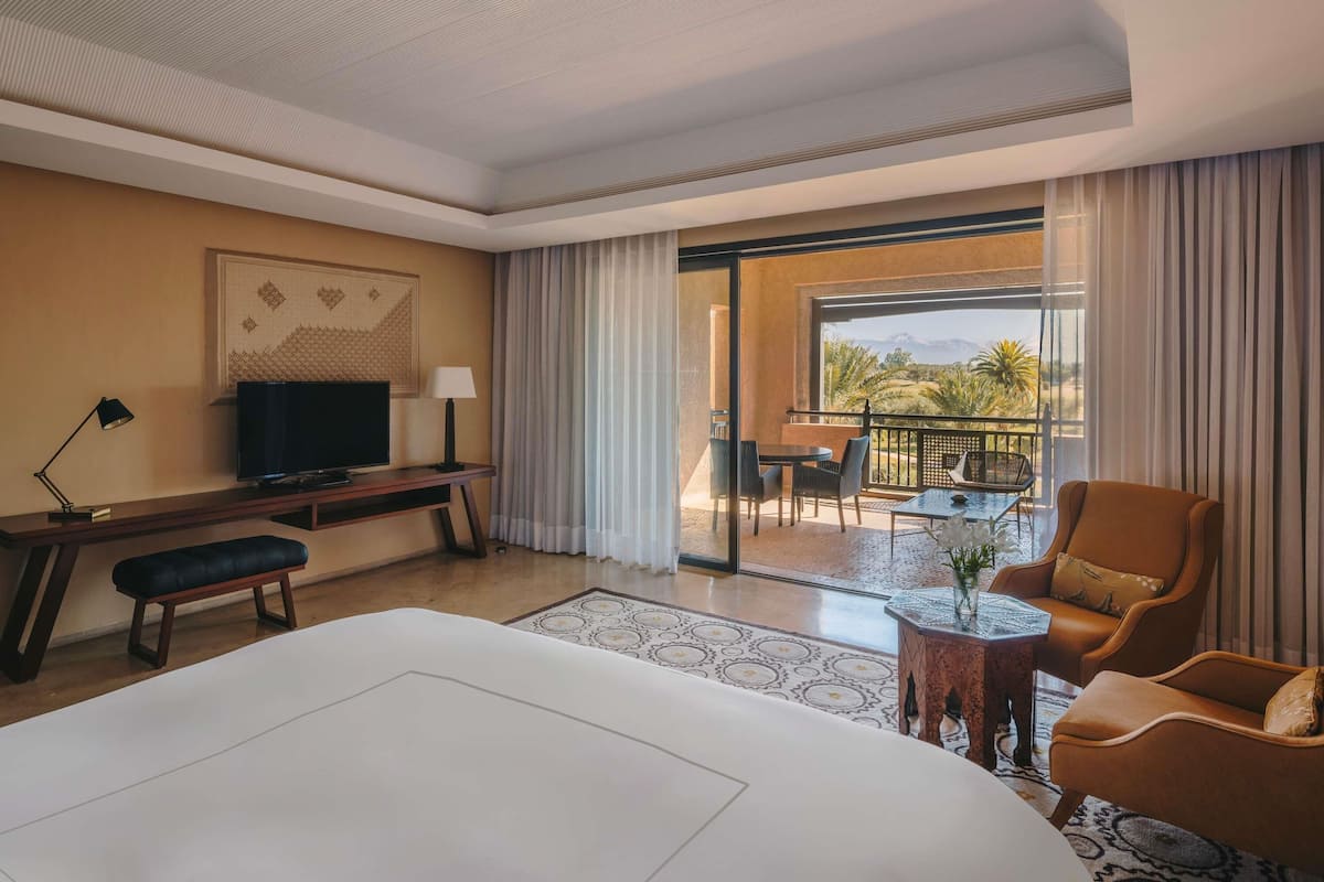 Une chambre d’hôtel avec un lit, un bureau avec une télévision, un balcon avec une table et des chaises, et une vue sur les palmiers.