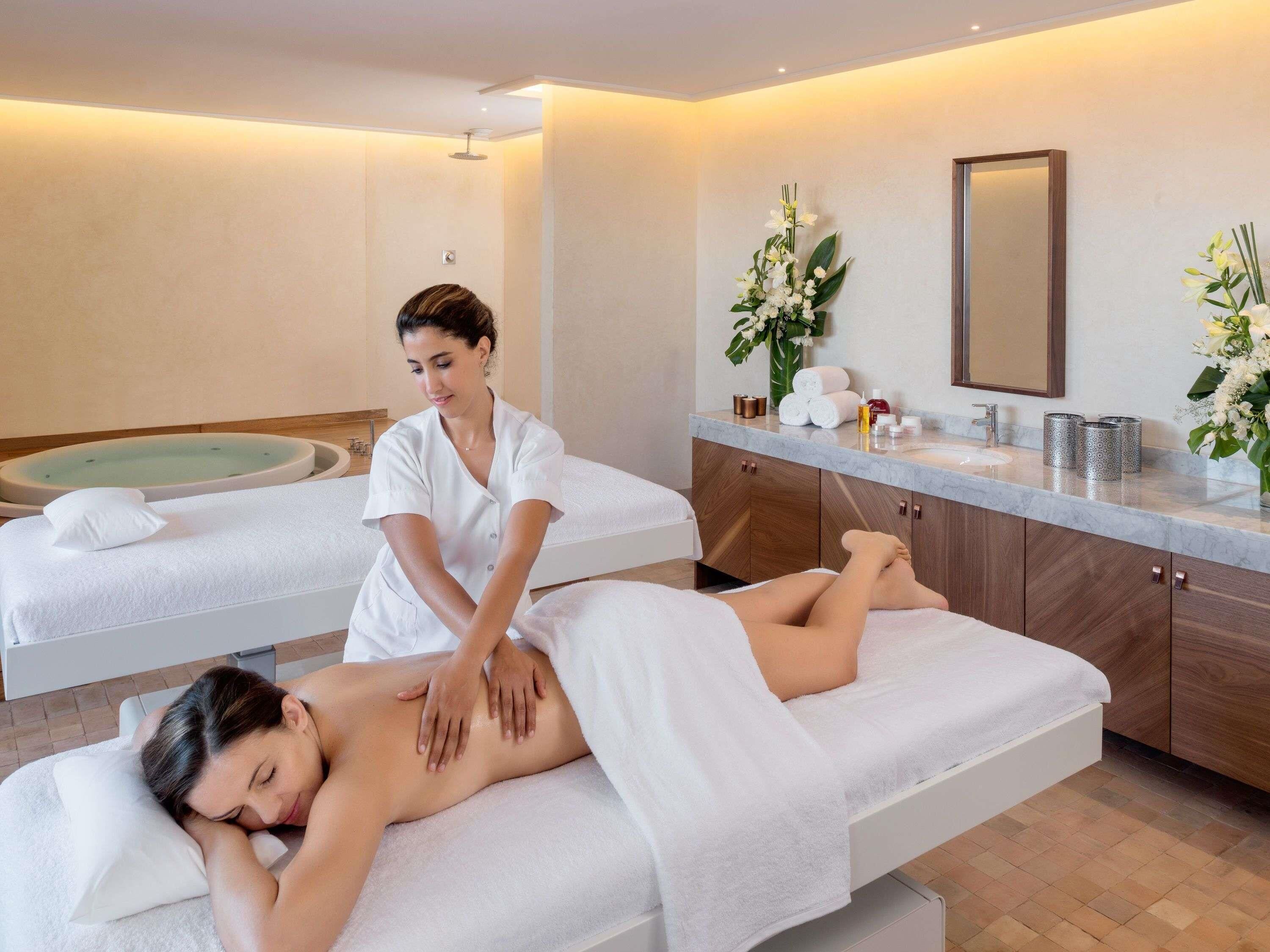 A spa room with two massage tables, a wooden cabinet with towels, and a mirror.