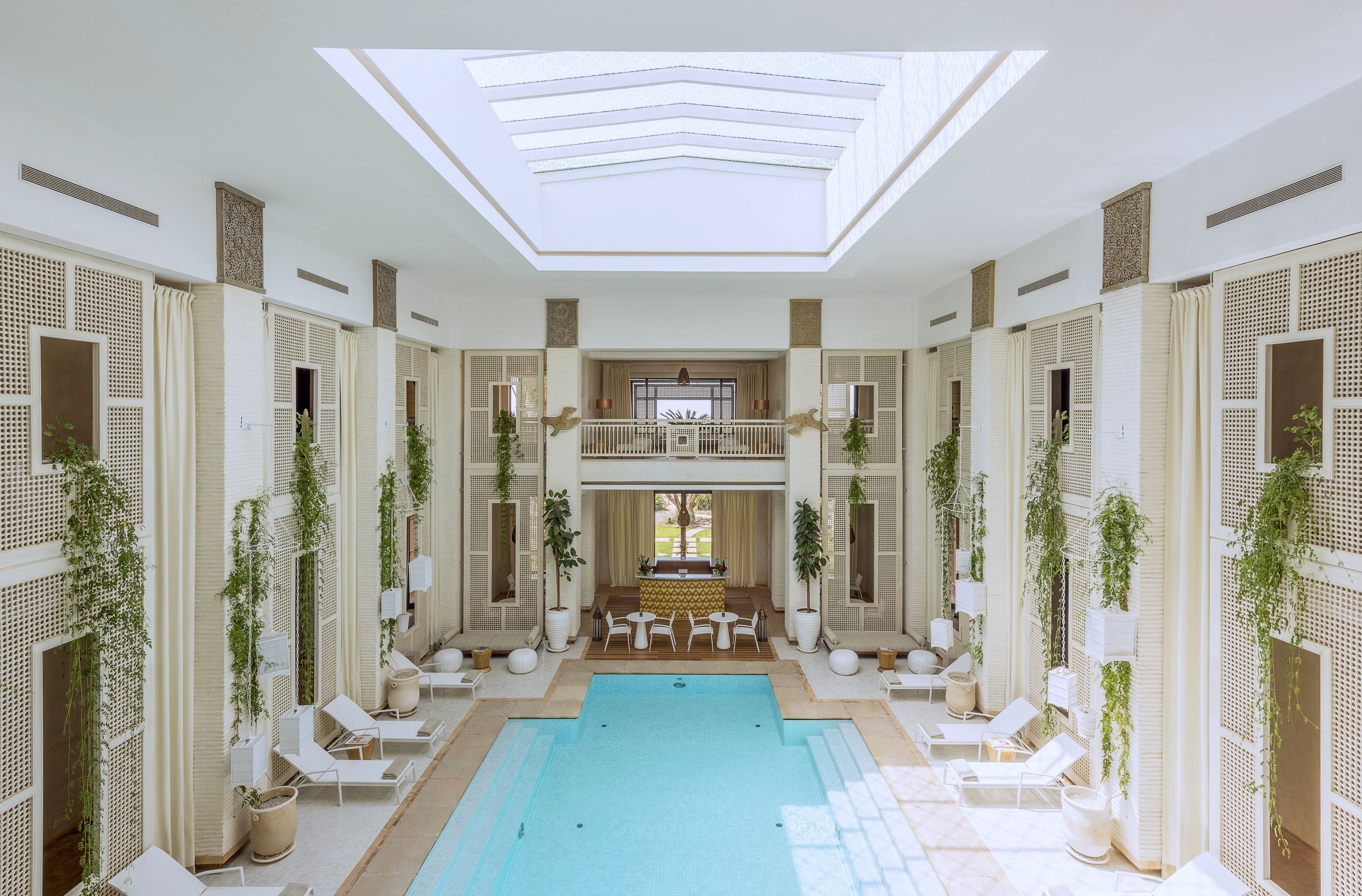 A spacious indoor swimming pool with a central seating area, surrounded by white lounge chairs and potted plants.