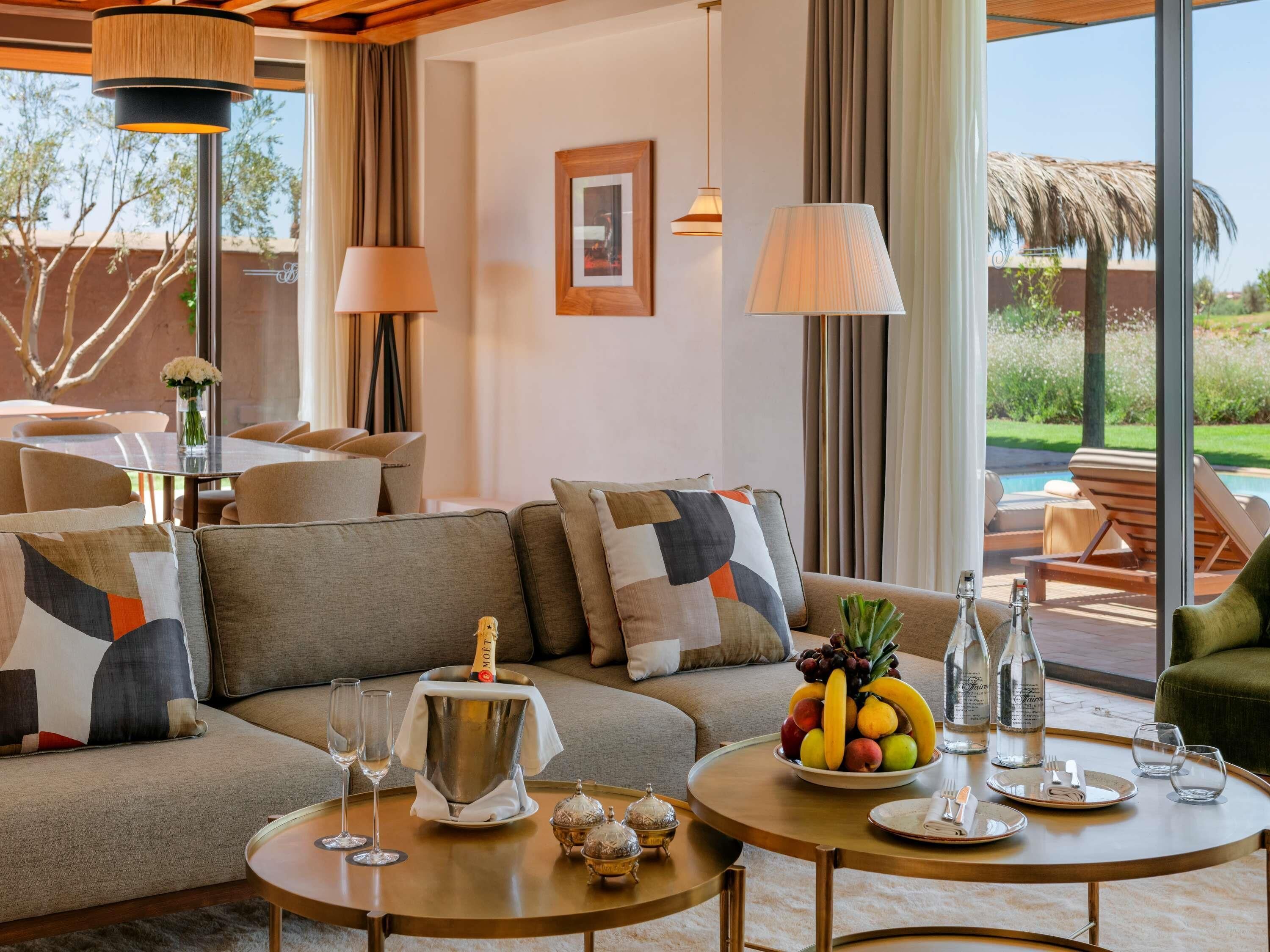 A modern living room with a sofa, a round coffee table, and a dining area with a fruit bowl and bottles.
